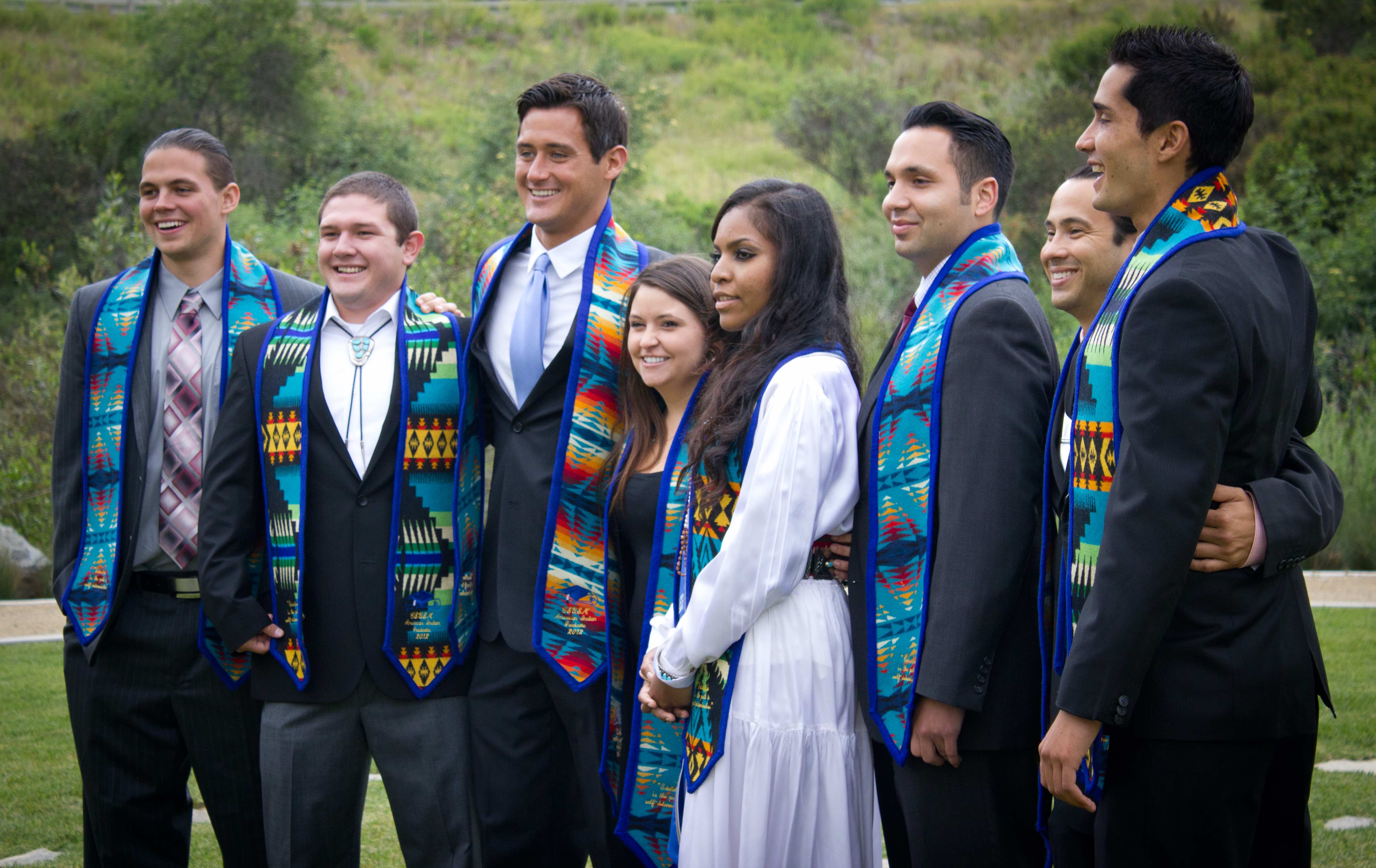 Group of American Indian graduates standing in a horizontal line wearing graduation stoles over their formal clothes, smiling at the camera, standing outside on green grass in front of a landscape of green trees and hills