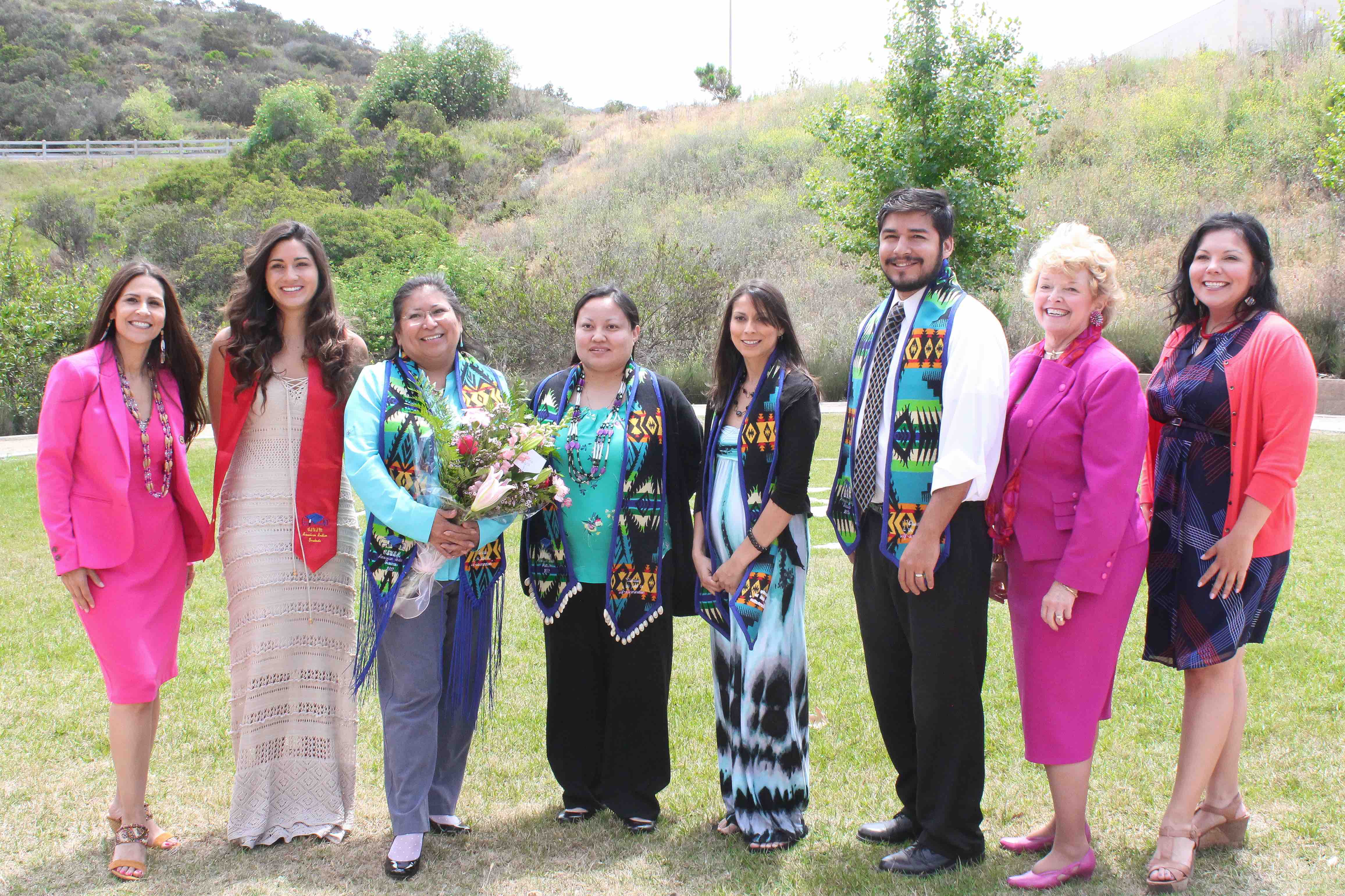 Group of American Indian graduates (as well as CSUSM faculty) standing in a horizontal line wearing graduation stoles over their clothes, smiling at the camera, standing outside on green grass in front of a landscape of green trees and hills