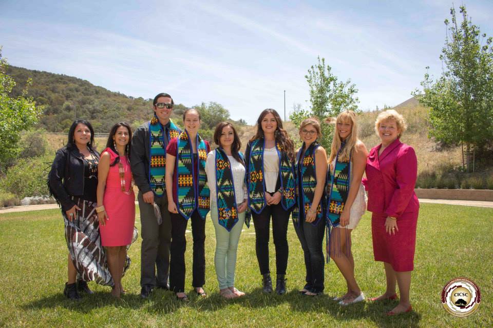 Group of American Indian graduates (as well as CSUSM faculty) standing in a horizontal line wearing graduation stoles over their clothes, smiling at the camera, standing outside on green grass in front of a landscape of green trees and hills, and a blue sky