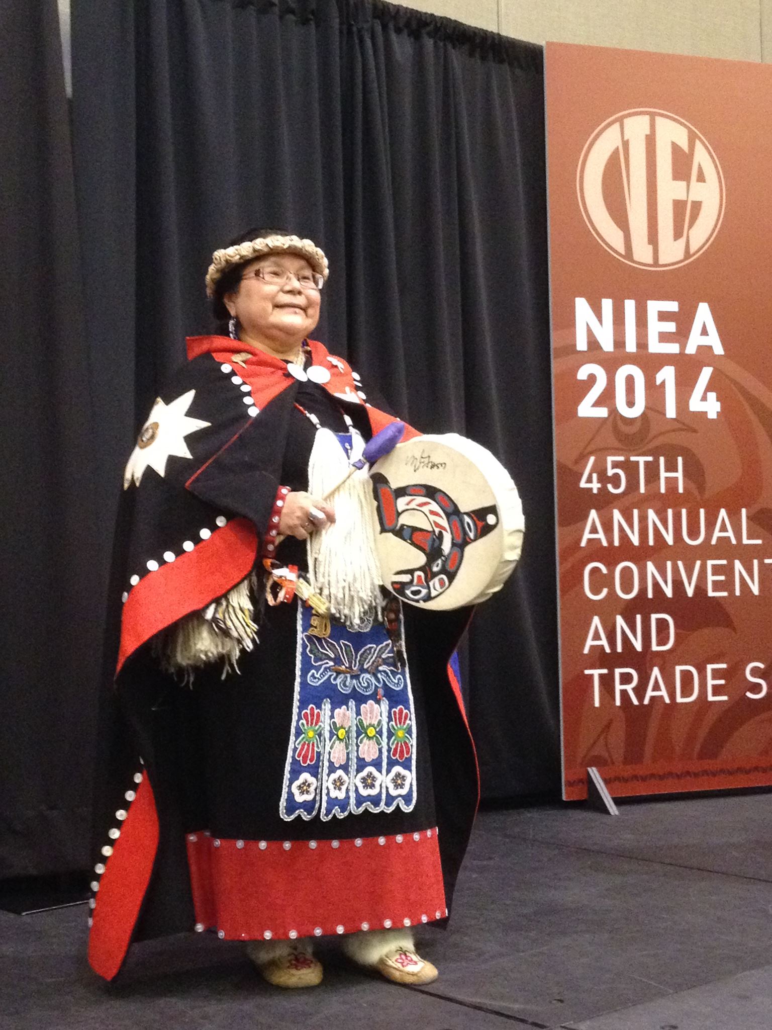 Alaskan Native Cultural Night at NIEA - a woman in traditional dress standing next to a sign that says, "NIEA 2014 45th Annual Convention and Trade Show"