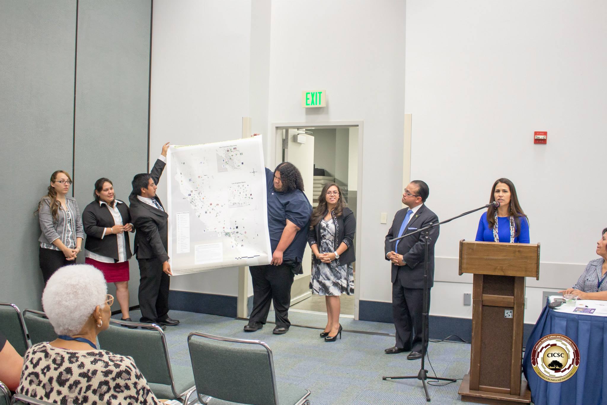 Dr. Joely Proudfit speaking at a podium and students from CSUSM's American Indian Student Alliance standing in front of the room, holding and standing around a large GIS research poster, as people in the audience and someone at a panel table next to Dr. Proudfit listen to the presentation