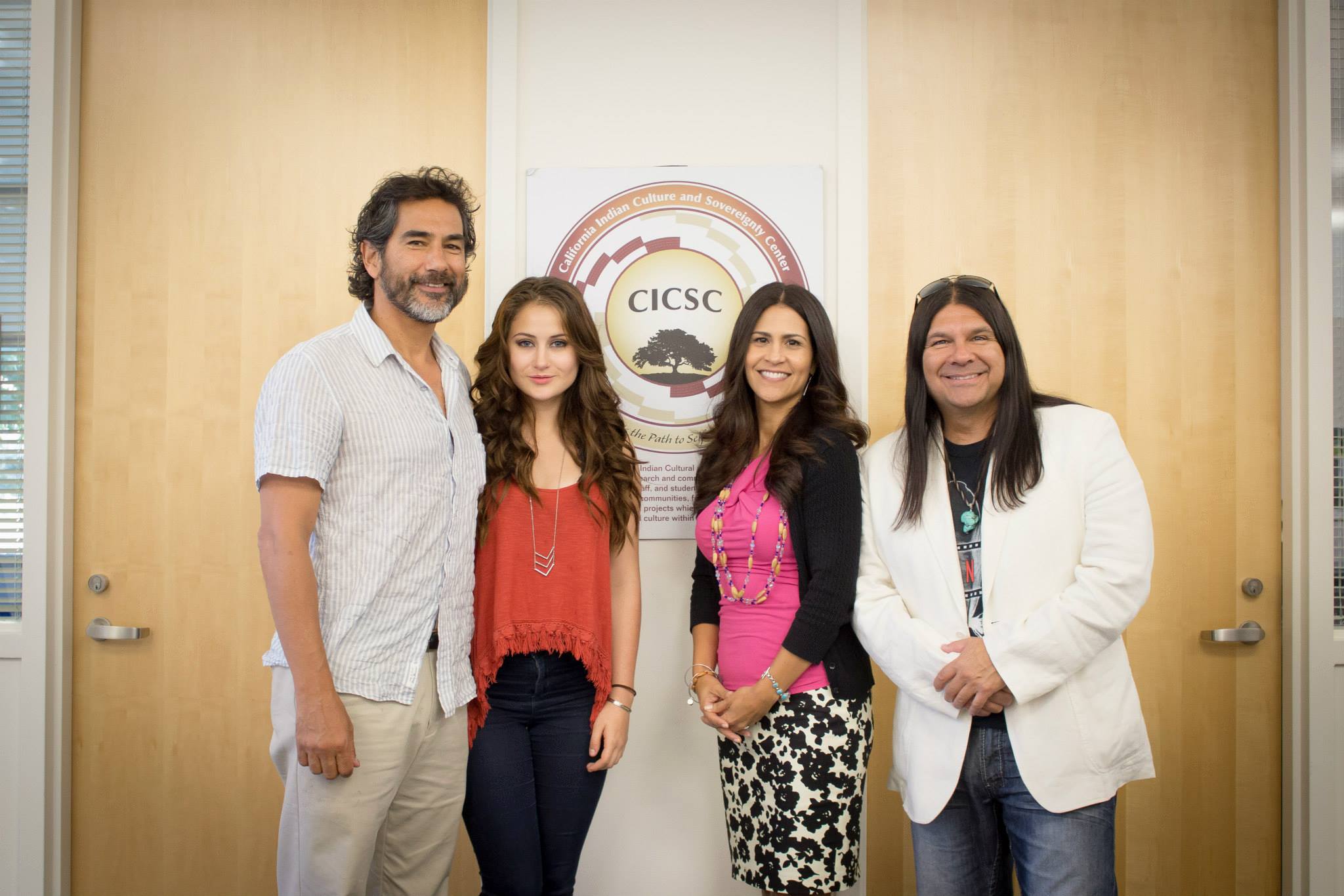 Bella King, Ruben CHATO Hinajosa, and Randy Vasquez standing with Joely Proudfit in front of a sign displaying the CICSC logo on a white wall in between two wooden doors in the CICSC center on campus