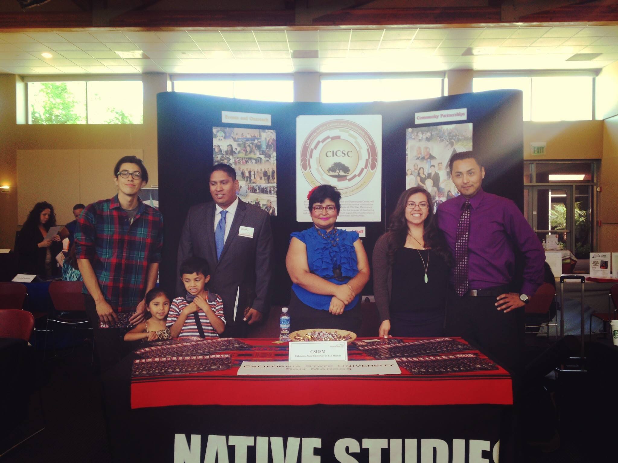 A group of people standing behind a table with various brochures and items on it, and a tablecloth that says "Native Studies" on the front; the people are also standing in front of a large tri-fold poster with a large CICSC logo on it as well as collages of pictures
