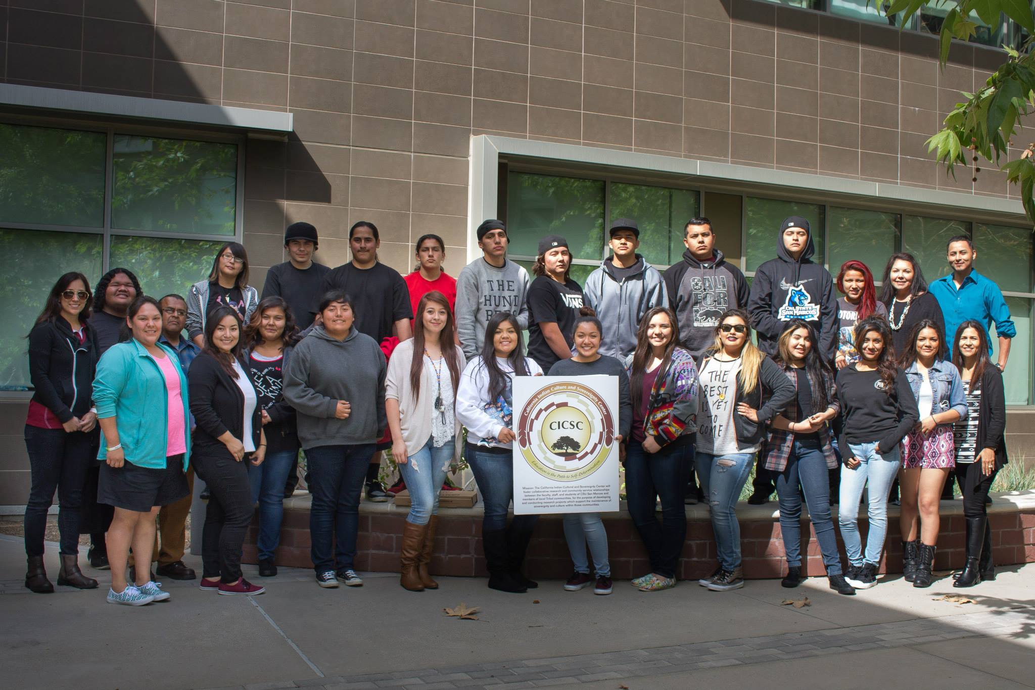 A large group of Noli Middle & High School students standing together in two rows outside of a building, and two people in the front row are holding a sign with the CICSC logo
