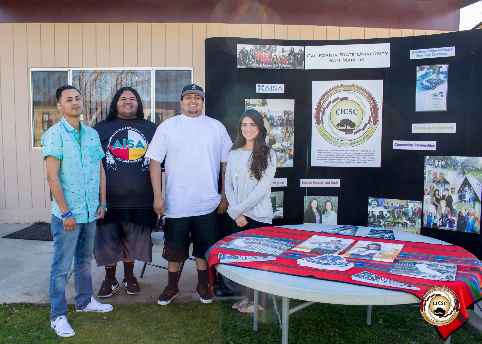 Four students from the CICSC and AISA standing outside in front of a table and poster board displaying information about the CICSC, AISA, and Native American community and events at CSUSM