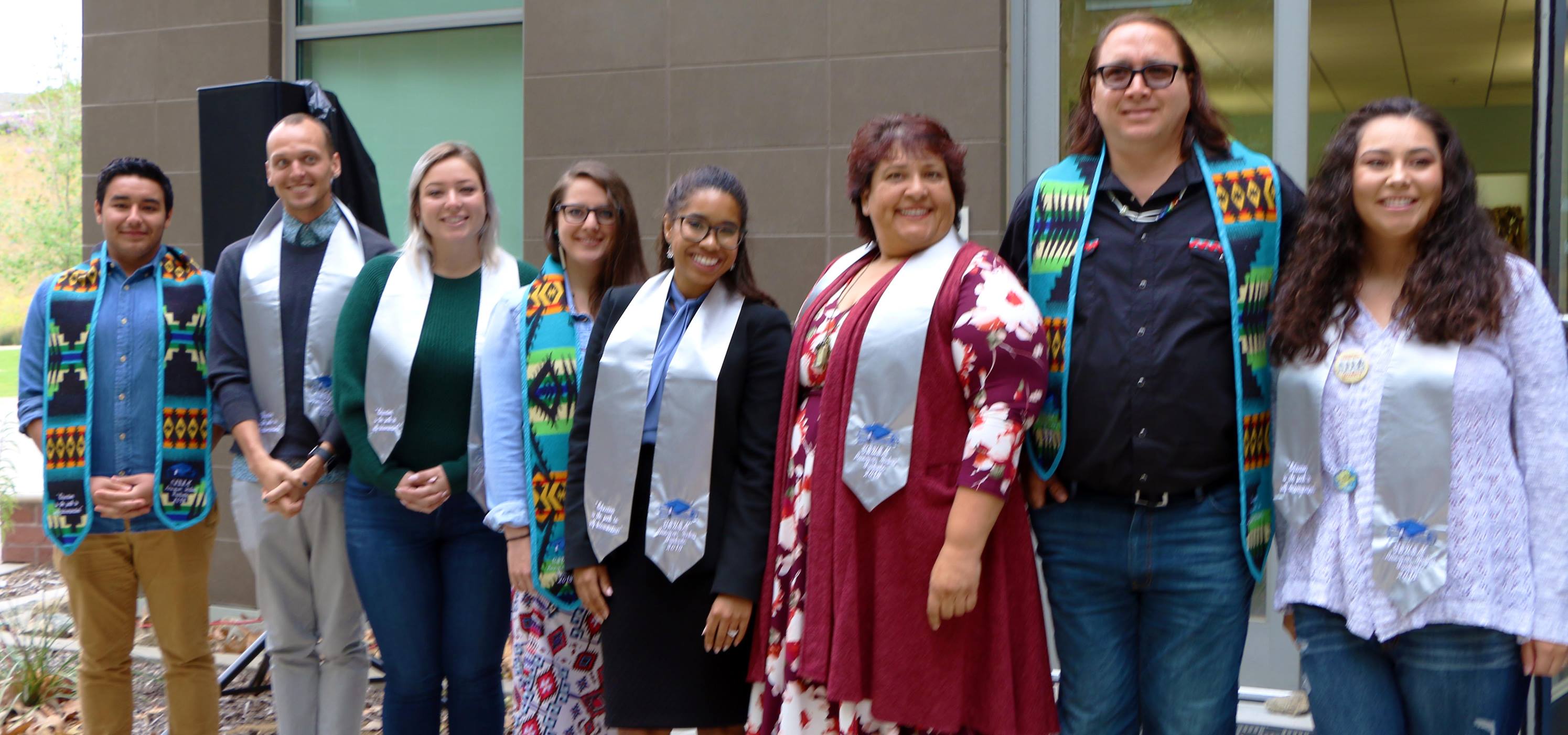Group of American Indian graduates standing in a horizontal line wearing graduation stoles over their clothes, smiling at the camera