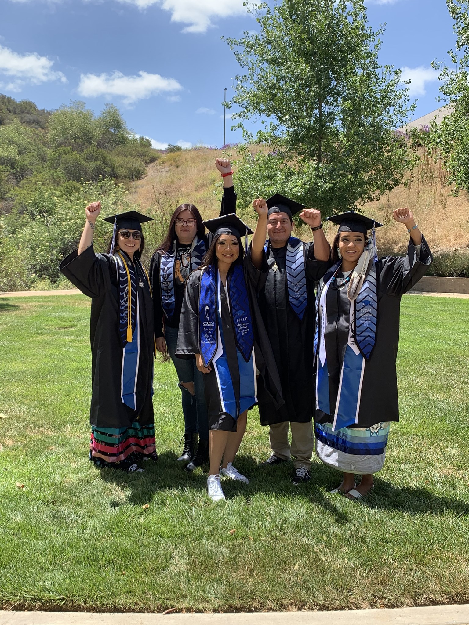 Group picture of American Indian graduates in their capa, gowns, and stoles,smiling and raising one fist