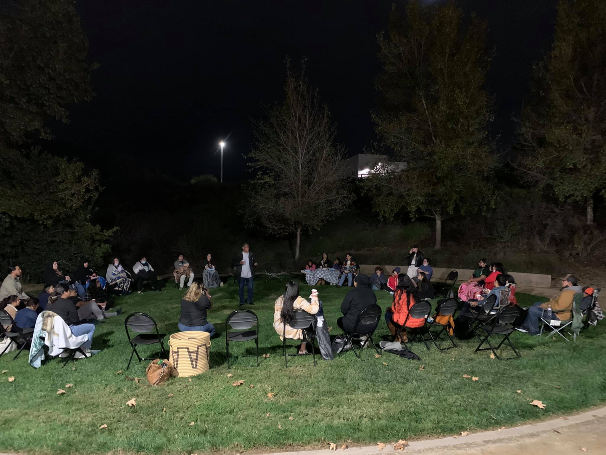 People sitting in a circle of black folding chairs placed on the grass around Dr. Stan Rodriguez as he stands in the center of the circle against the black nighttime sky