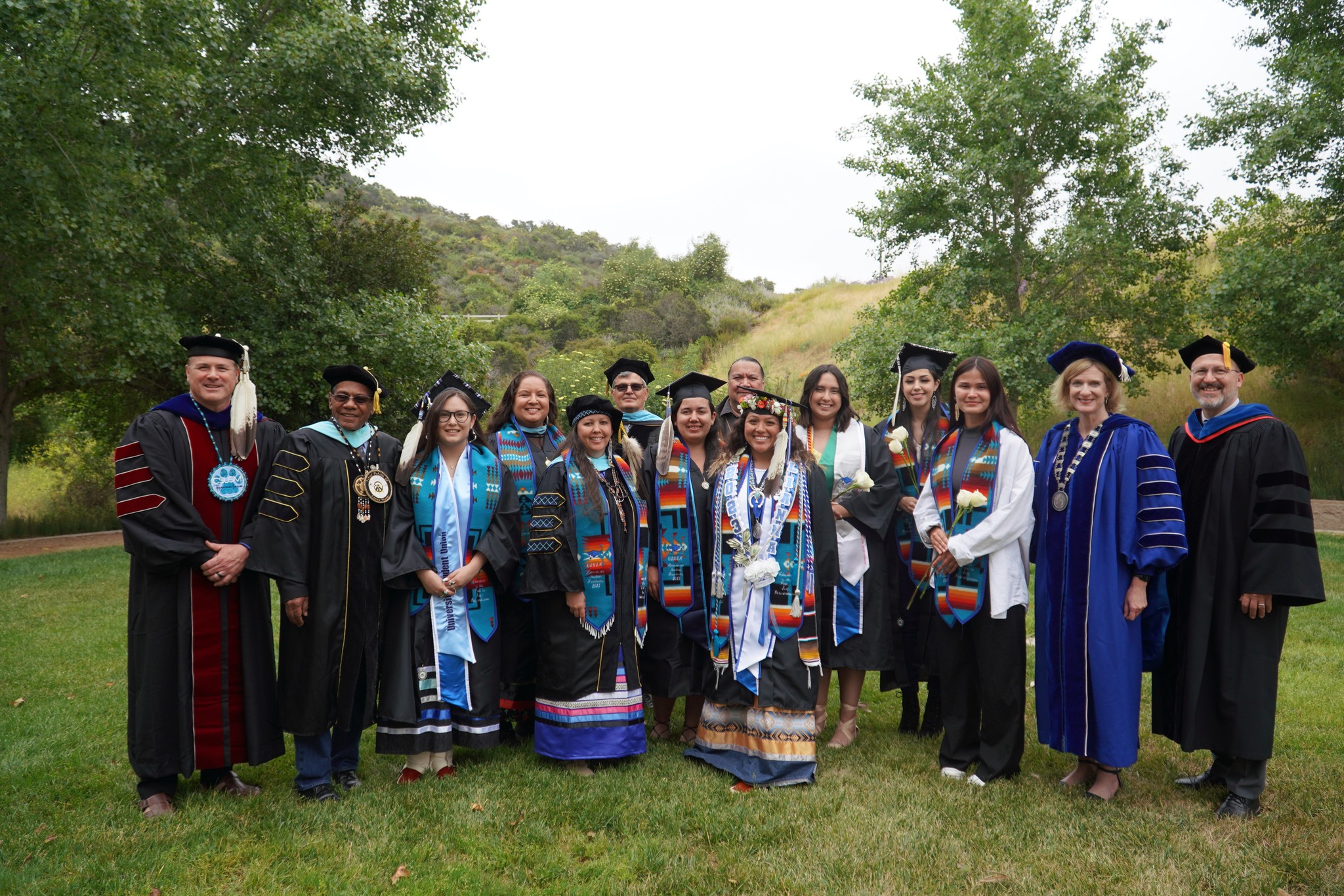 Group picture of American Indian graduates and faculty in their caps, gowns, and stoles, standing together outside in a green landscape of grass with trees and small hills in the background
