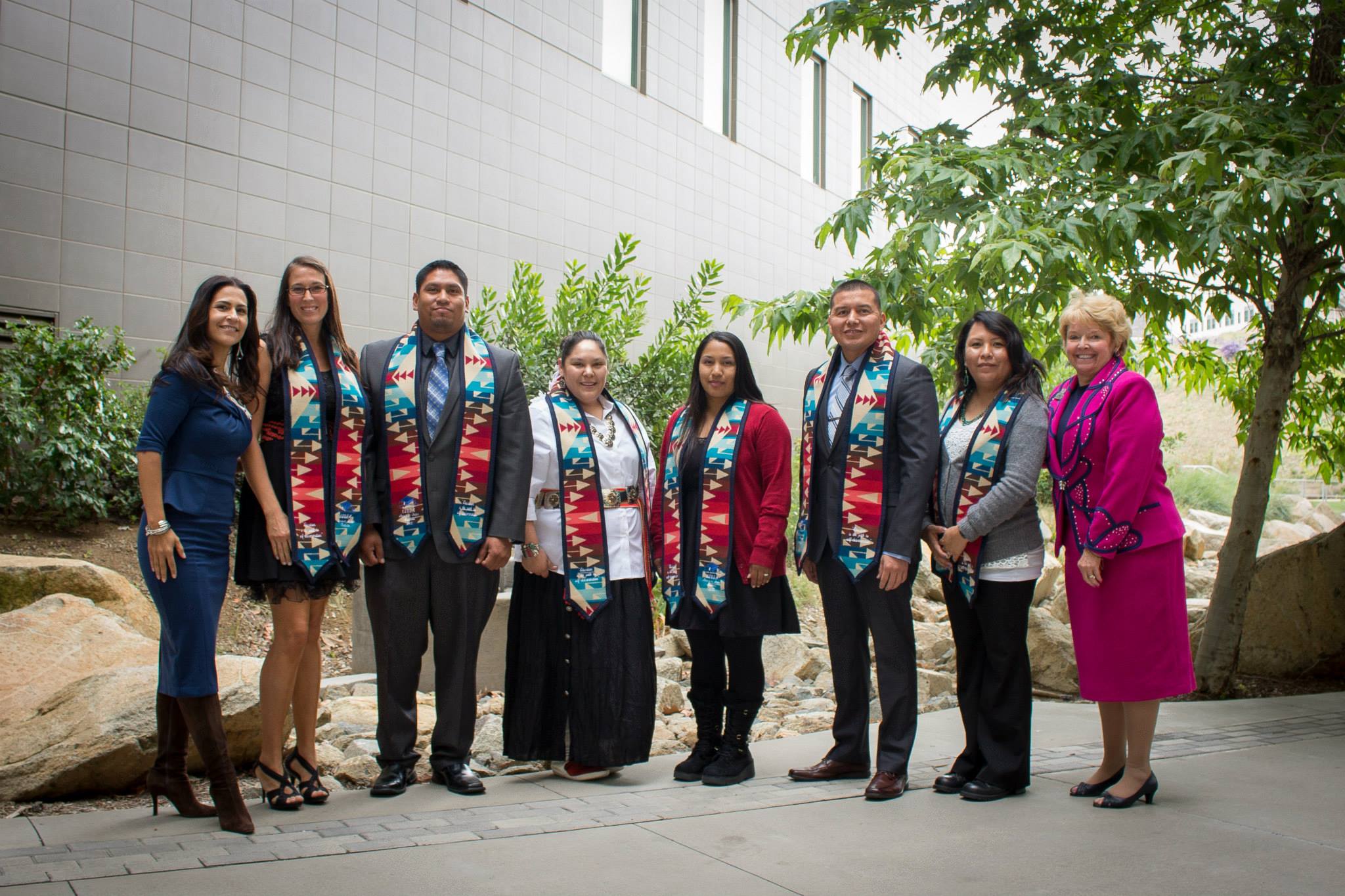 Group of American Indian graduates (as well as CICSC Director Dr. Joely Proudfit and CSUSM President Karen Haynes on either end) standing in a horizontal line wearing graduation stoles over their clothes, smiling at the camera, standing outside in front of greenery, rocks, and a white building wall