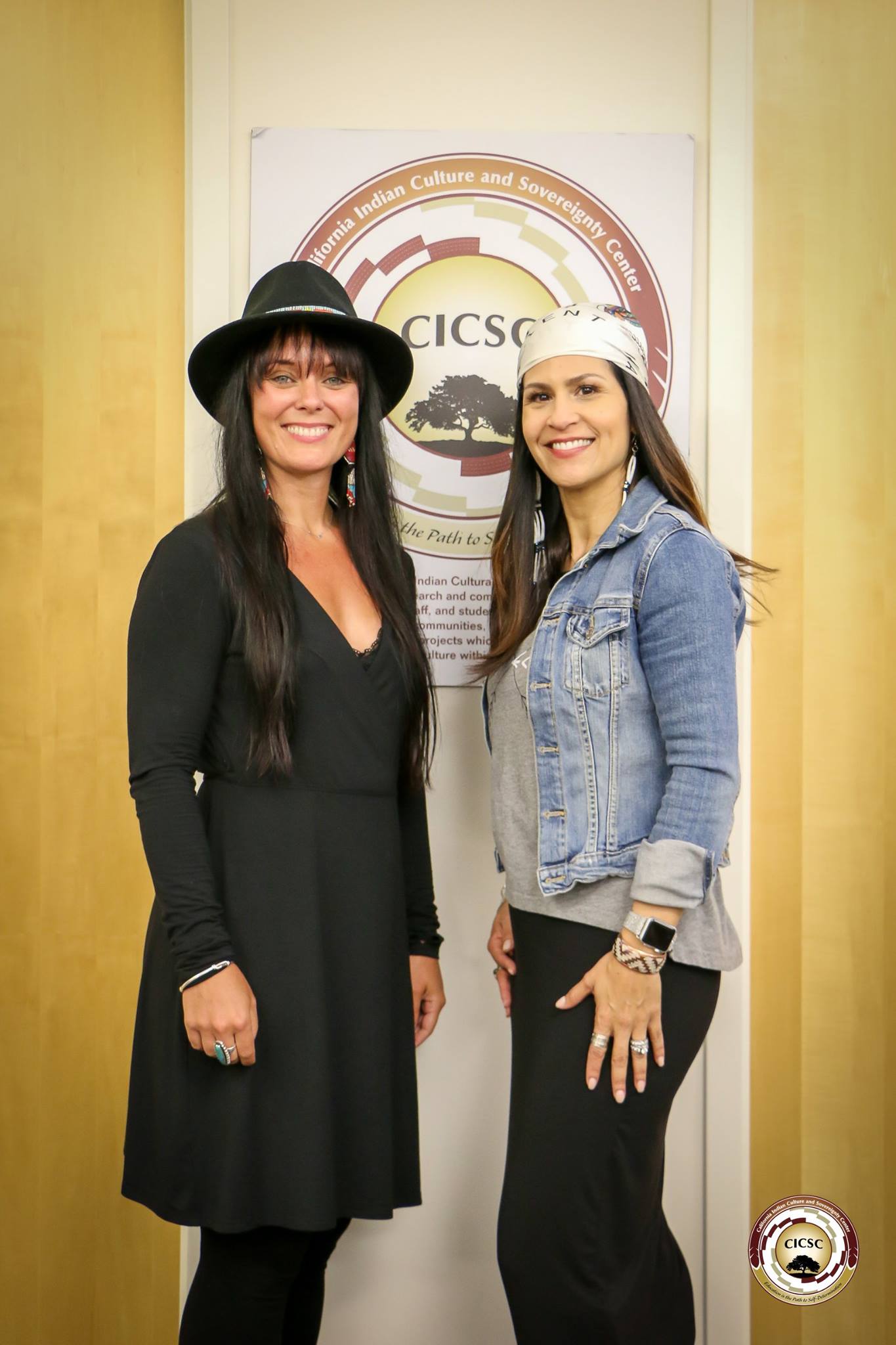 Director Michelle Latimer and CICSC Director Joely Proudfit standing next to one another in front of a CICSC logo sign hanging on a wall