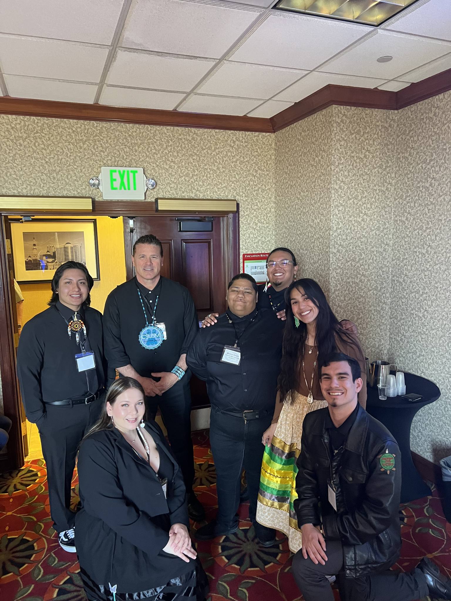 A group of students with Dr. Eric Tippeconnic, gathered together near the doors in the corner of a room, everyone wearing all-black formal clothing with one person wearing a brown shirt and a ribbon skirt with green, gold, brown, and black ribbons.  