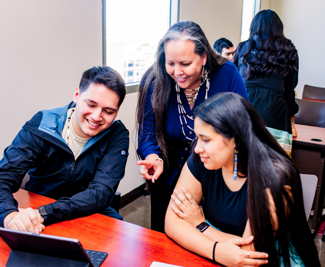 Lecturer Maillet standing in between two students seated at a table in a classroom, and leaning forward to point something out on an iPad with a keyboard to them