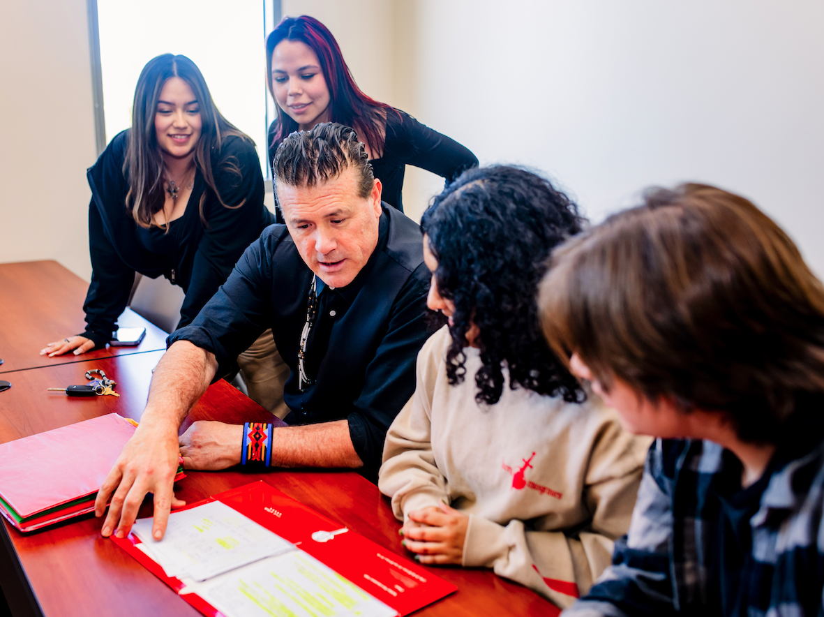 Eric Tippeconnic sitting at a table surrounded by students as he leans over a folder to explain something to everyone